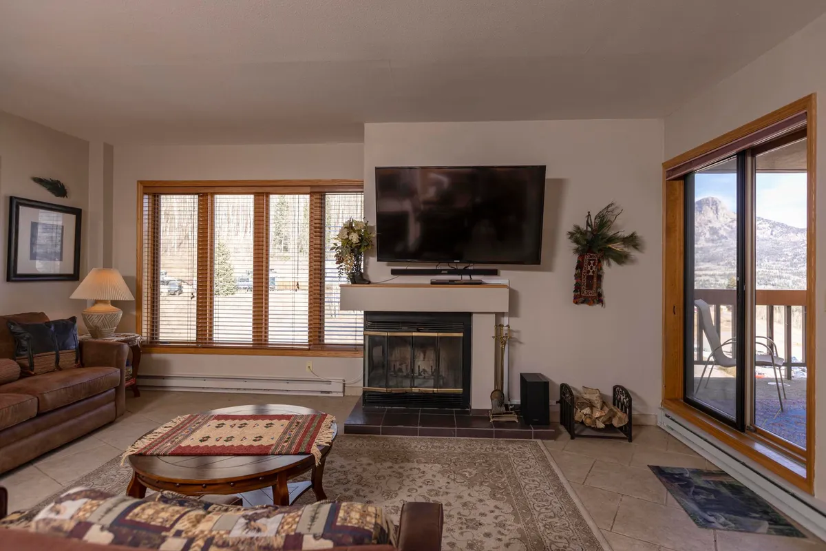 A warm, inviting living room featuring a prominent wood-burning fireplace, large windows letting in natural light, a plush sofa, coffee table, and television. The seating is grouped for easy conversation and relaxation, evoking a welcoming atmosphere ideal for gathering after a day on the slopes.