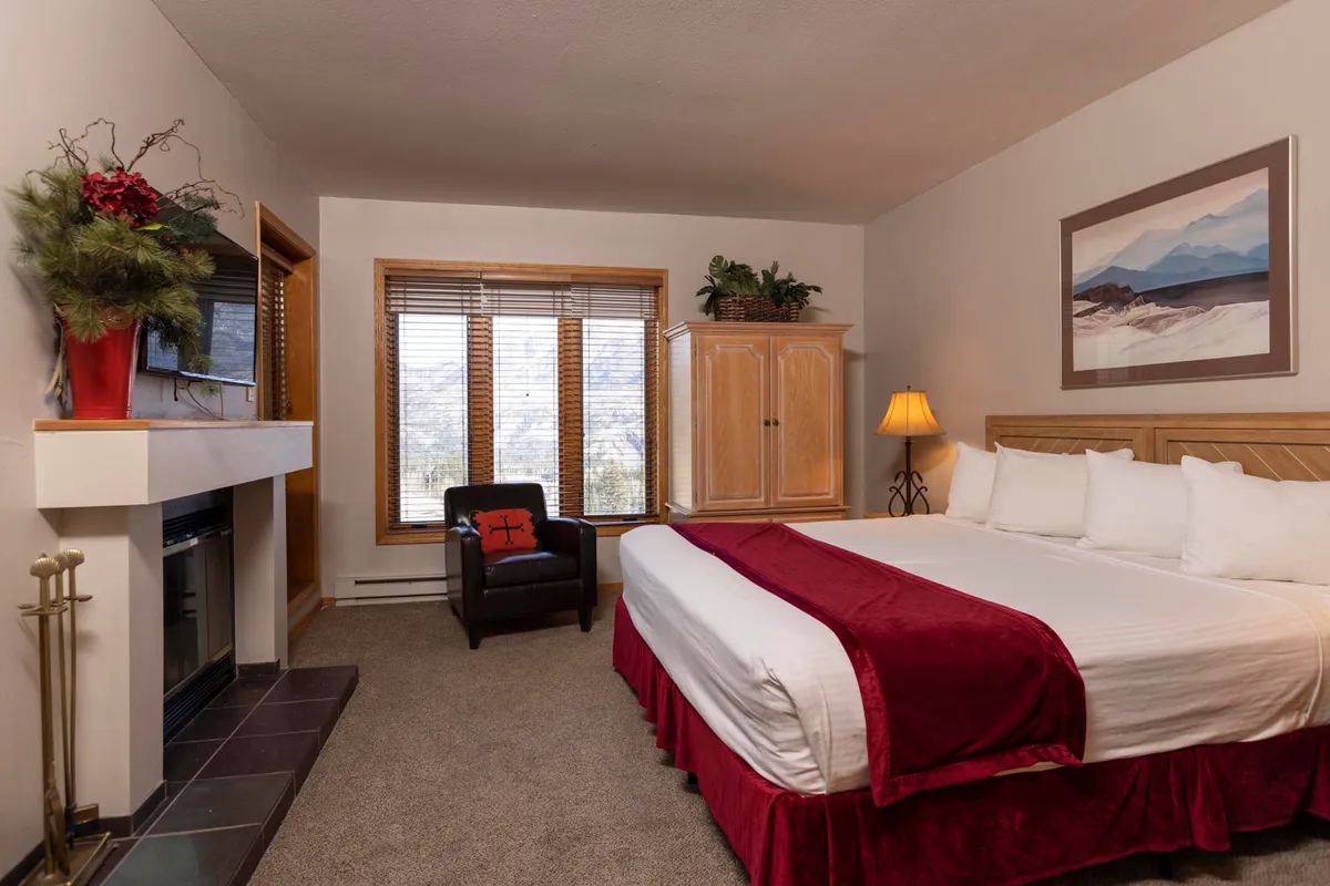 A cozy bedroom with an inviting bed adorned with red accents, a fireplace for warmth, and a large window that frames a scenic view. Warm wooden cabinetry and tasteful artwork add personal touches, making the space feel both luxurious and homey.