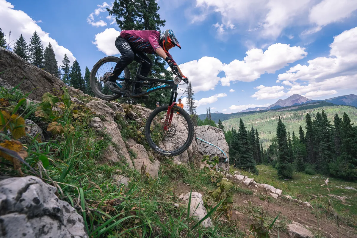A mountain biker rides along rocky terrain with lush greenery and distant mountain peaks forming a dramatic backdrop. The scene is alive with the thrill of adventure, capturing the essence of outdoor exploration in the San Juan Mountains.