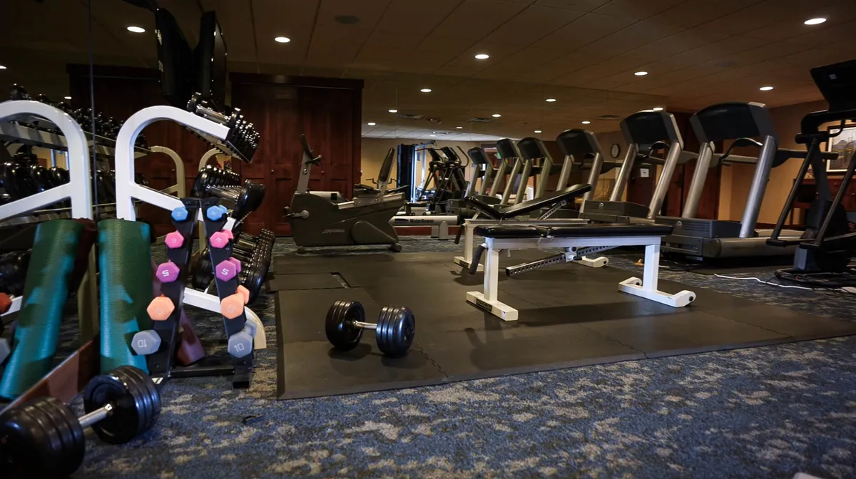 An inviting, well-equipped fitness center featuring rows of dumbbells, treadmills, a weight bench, and exercise mats. The bright, motivational space is designed for guests to recharge and maintain wellness routines, with ample room for both solo and group workouts.