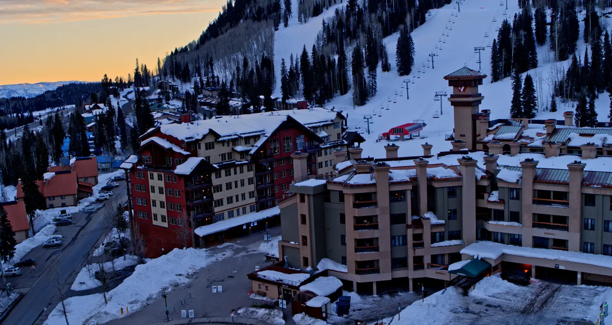 This exterior image captures snow-covered resort buildings nestled among tree-lined mountains, with ski lifts ascending the slopes beneath a soft twilight sky. The scene exudes a cozy yet adventurous mountain resort atmosphere.
