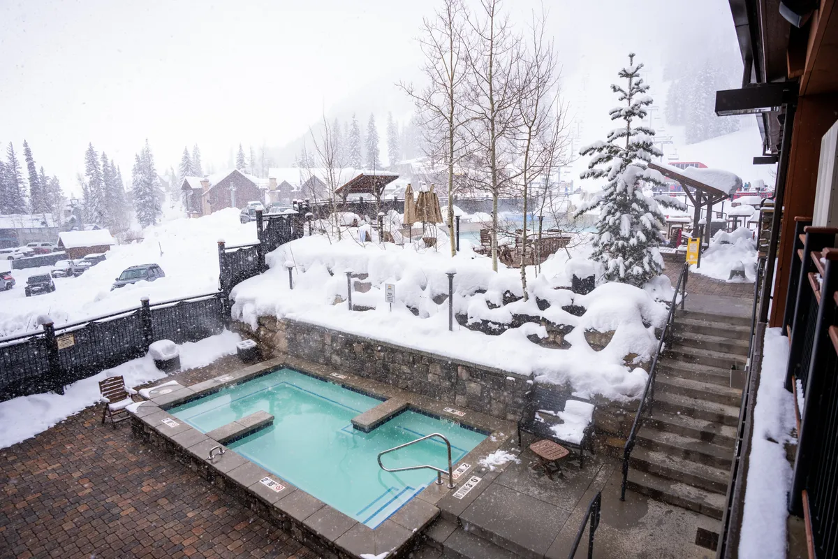 An outdoor hot tub steams invitingly amid a snowy landscape, surrounded by trees and a staircase leading to the tub. The setting is serene yet invigorating, promising relaxation after a day spent exploring the mountains.
