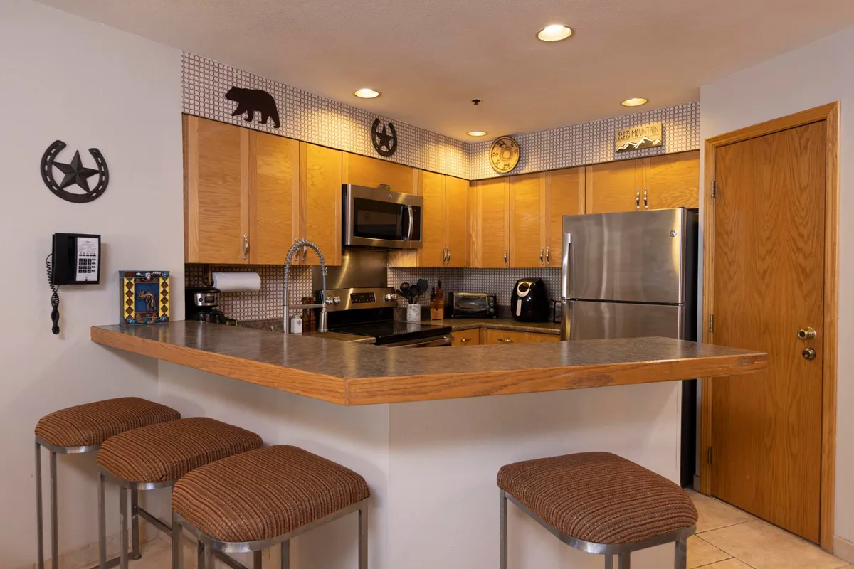 This image showcases a central kitchen island with stools set up for casual dining, surrounded by modern appliances and decorative wall art. The warm lighting and inviting atmosphere suggest a space perfect for gathering, cooking together, and sharing meals.