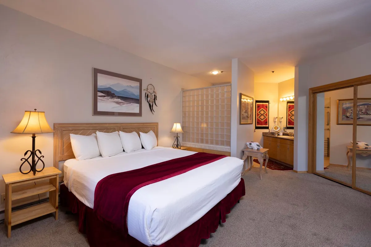 A bedroom with crisp white linens accented by burgundy, ambient lighting from bedside lamps, and tasteful wall art. The open layout offers a glimpse into a well-appointed bathroom area, underscoring the convenience and luxury of en suite facilities.