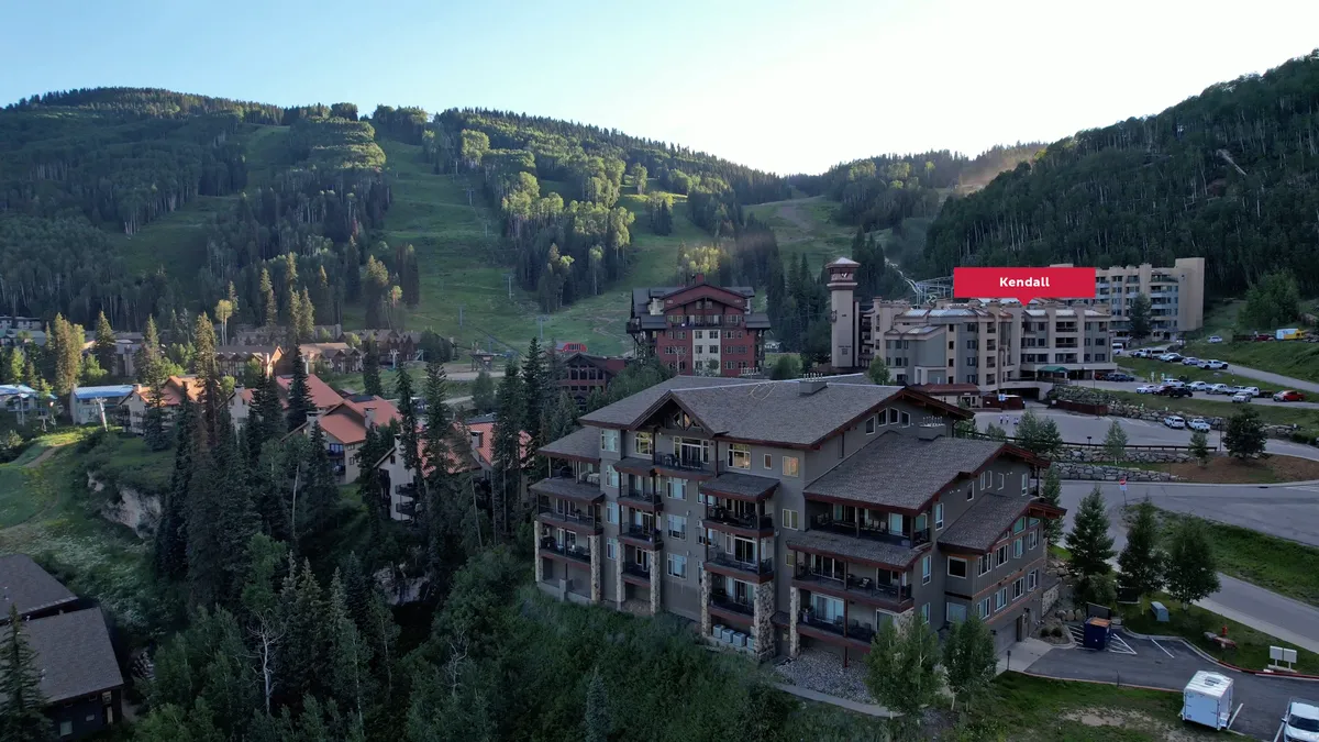 A panoramic view of the resort buildings nestled among dense trees with the dramatic mountain range rising behind. The scene conveys a sense of community, adventure, and proximity to outdoor activities.