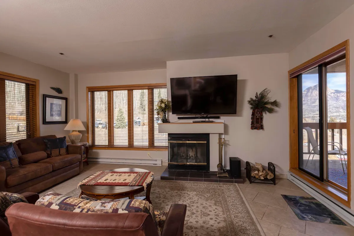 The image showcases a cozy, tastefully furnished living area featuring a plush seating arrangement centered around a wood-burning fireplace. Large windows flood the room with natural light and offer glimpses of the surrounding mountain scenery, creating an inviting retreat perfect for relaxing after a day of outdoor adventure.