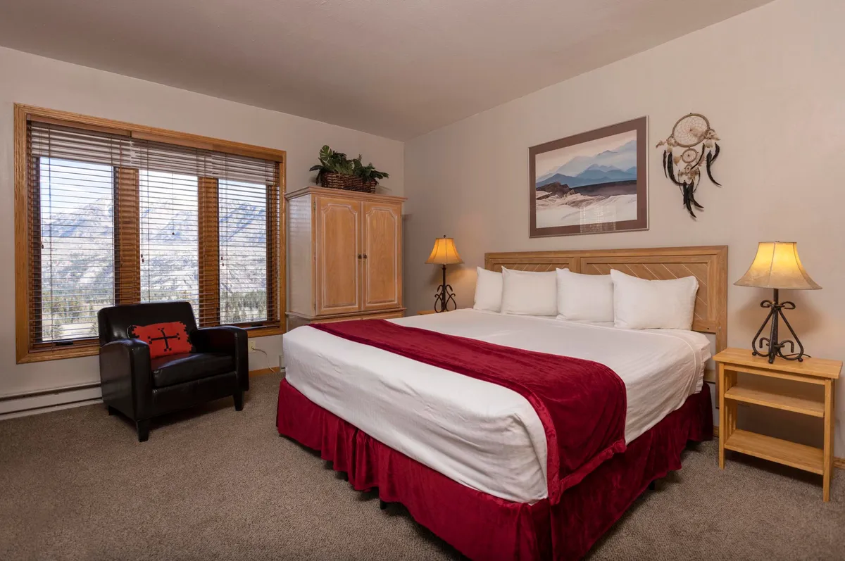 A spacious bedroom featuring a large, comfortable bed dressed in plush bedding, flanked by windows that let in natural light and offer stunning mountain views. An armchair sits in the corner beside a nightstand with a softly glowing lamp, creating a tranquil and inviting space.