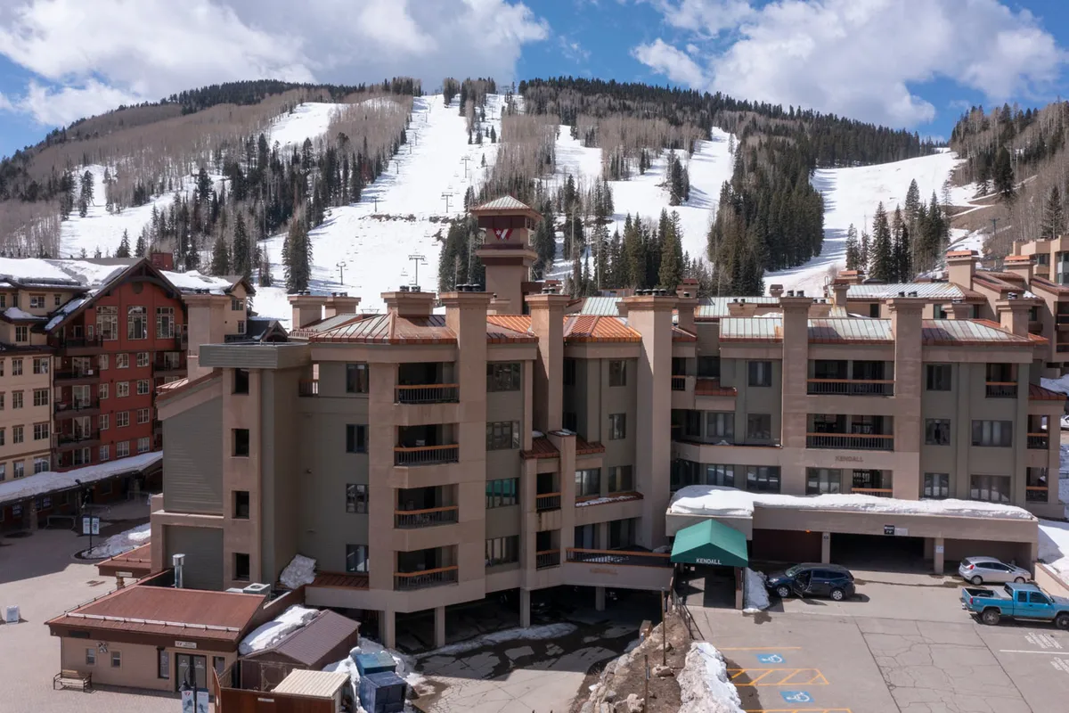The Kendall building stands at the base of snow-covered slopes, with its inviting architecture framed by breathtaking mountain views and a lively resort atmosphere. Guests and visitors can be seen enjoying the winter landscape near the parking area.