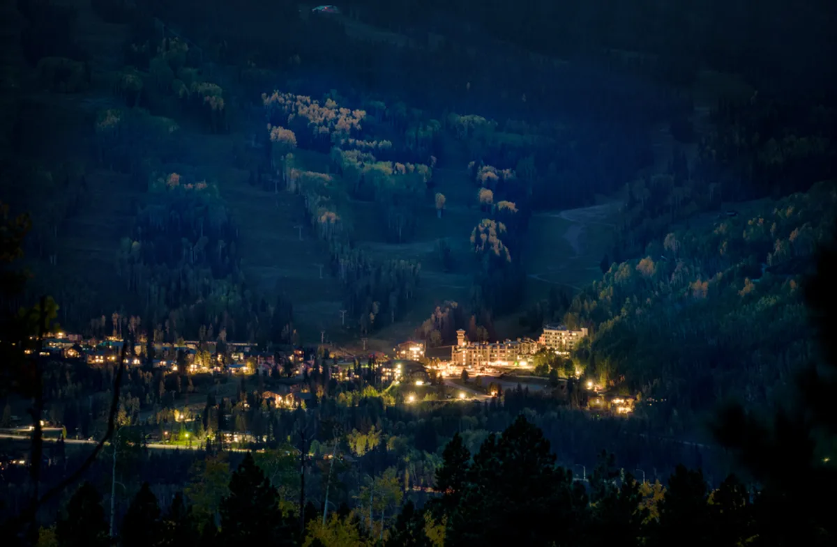 An exterior nighttime scene showing warmly illuminated resort buildings surrounded by forest and dramatic mountain landscapes. The sky is dark, perfect for stargazing. The ambiance is magical, cozy, and inviting—ideal for evening strolls or quiet moments together.
