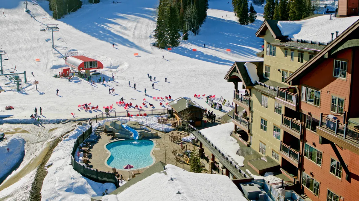 An exterior view of the resort’s main buildings with the pool area visible in the foreground, snow-dusted ski slopes beyond, and a ski lift nearby. The image captures both relaxation (pool) and vibrant activity (skiing, après-ski gatherings) within a lively village environment.